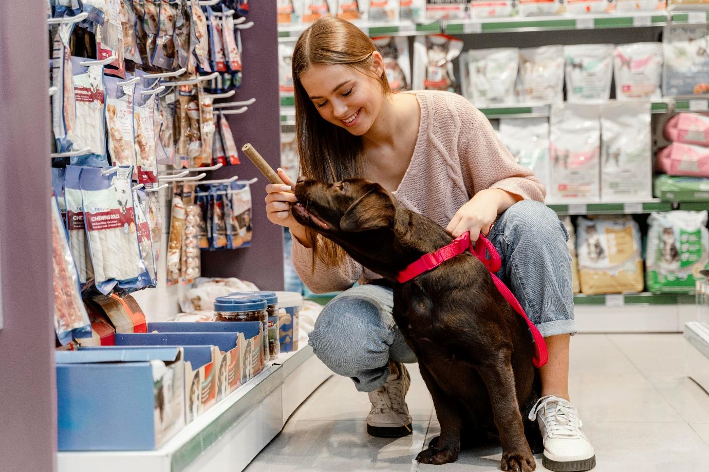 A woman crouches next to her dog in a pet store. The dog really wants the treat the woman has in her hand.